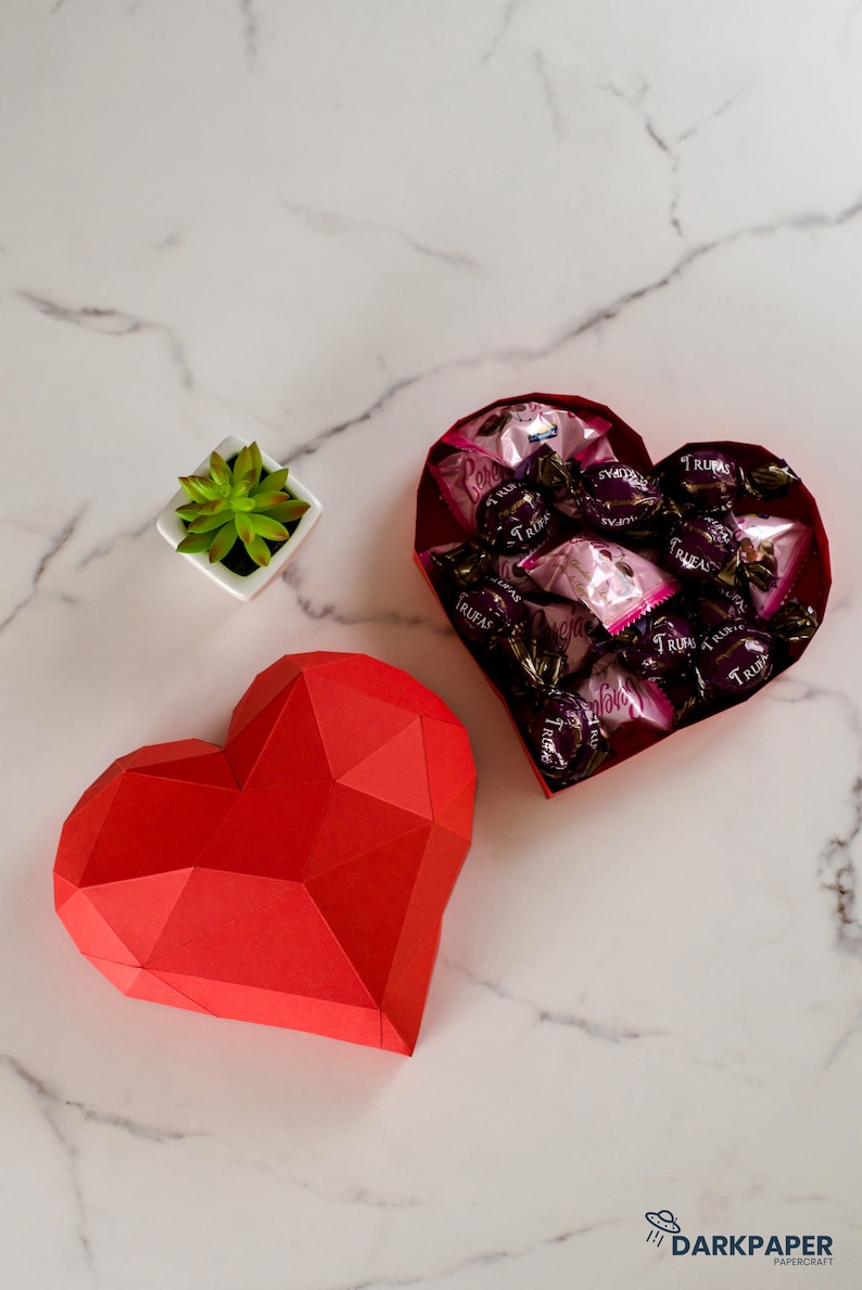 May include: A red paper heart-shaped box with a geometric design. The box is open and filled with small, individually wrapped chocolates. The chocolates are wrapped in pink and silver foil and have the word "Trufas" printed on them.