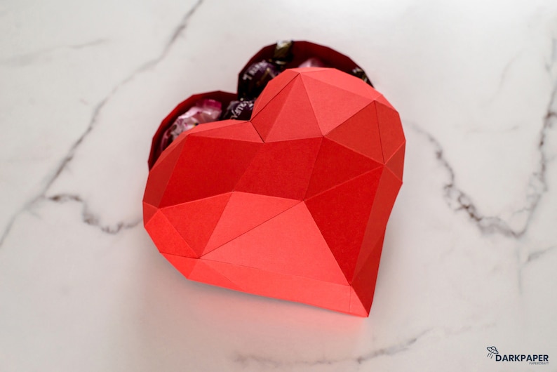 May include: A red paper heart-shaped box with a geometric design. The box is open and filled with small, wrapped chocolates.