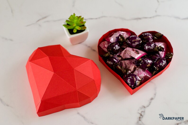 May include: A red paper heart-shaped box with a geometric design. The box is open and filled with wrapped chocolates. A small potted succulent plant is in the background.