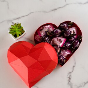 May include: A red paper heart-shaped box with a geometric design is open and filled with small chocolate candies wrapped in pink and gold foil. The candies are labeled "Trufas".