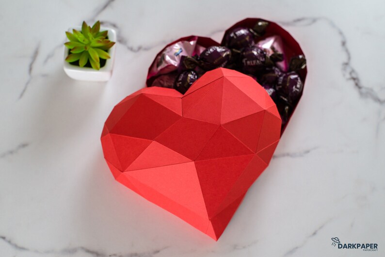 May include: A red origami heart-shaped box with a geometric design, open to reveal a collection of dark chocolate candies inside.