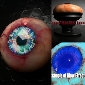May include: A realistic eyeball phone grip featuring a detailed blue and green iris with red veins on the sclera. The device includes a black circular base and displays a bright blue glow effect in low light conditions with visible text overlays.