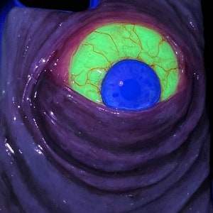 May include: A phone case with a 3D design of a glowing green eye with a blue pupil. The eye is surrounded by a dark purple, textured material.
