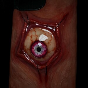 May include: A phone case with a realistic, grotesque design featuring a large, fleshy eye with a pink iris and a black pupil. The eye is surrounded by stitched flesh and appears to be bulging out of the case.