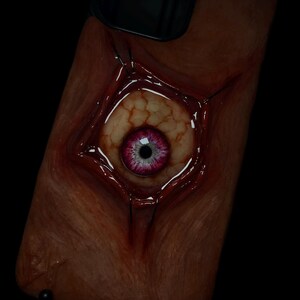May include: A phone case with a realistic, grotesque design. A fleshy, bloody, human-like eye is depicted on the case, with a pink iris and black pupil.