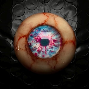 Könnte beinhalten: Ein rundes, realistisches Augapfel-Ornament mit einer blassen, fleischfarbenen Sklera und markanten roten Gefäßdetails. Die Iris ist eine lebendige Mischung aus Blau- und Magentatönen, die eine dunkle, kreisförmige Pupille umgibt.