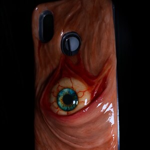 May include: A phone case with a fleshy, textured design featuring a large, blue eye with red veins. The case is made to look like a human eye.