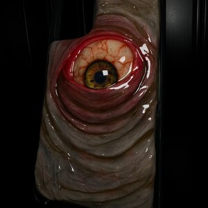May include: A close-up of a phone case with a realistic, grotesque eye design. The eye is red and bloodshot with a yellow iris and black pupil. The case is made of a textured, fleshy material.