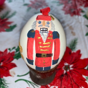 May include: A hand-painted egg ornament depicting a nutcracker in a red and blue uniform with gold accents. The egg is white with a brown base.