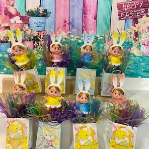 May include: Nine small Easter bunny figurines with bunny ears, each holding a small gift bag. The gift bags are decorated with Easter themes, including chicks, baskets, and eggs. The figurines are surrounded by green and purple Easter grass. The background is a pink and blue striped wall with a sign that says "Happy Easter!"