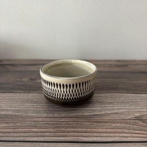 May include: A small, white ceramic bowl with a brown and white geometric pattern. The bowl is round and has a slightly flared rim.