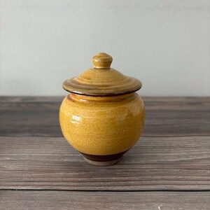 May include: A small, yellow ceramic jar with a lid. The jar has a brown glaze and a slightly textured surface.