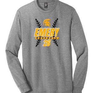 May include: A long-sleeved, heather gray shirt with the text "EMERY BASEBALL 26" in yellow and black. The design includes a yellow helmet graphic and black baseball stitching. The shirt has a crew neck and long sleeves.