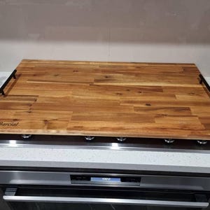 May include: A rectangular wooden chopping board with a natural wood grain pattern. It has black handles and the text "Team Barcial". The board is approximately 61 cm long.