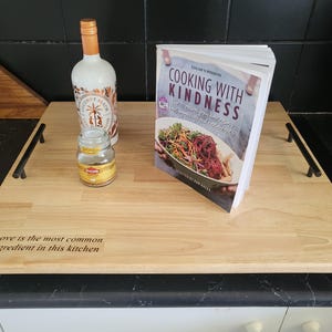 May include: A wooden serving tray with black handles. The tray has a black border and a light wood surface. The tray has the text "Love is the most common ingredient in this kitchen" engraved on the surface. A cookbook titled "Cooking with Kindness" is on the tray. A clear glass jar and a bottle of liquor are also on the tray.