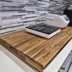May include: A wooden chopping board with a handle and the engraved word "Payuyo". A white and black appliance and a rice cooker sit on top of the board. The board is made of brown wood.