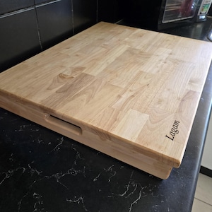 May include: A large, rectangular wooden chopping board with a light brown finish. The board has a raised edge and a handle on the side. The words "Lagam" are engraved on the board.