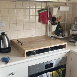 May include: A light-coloured wooden stove cover with black handles. The cover sits over a black hob. Kitchen accessories, including oven mitts, utensils, and a kettle, are visible in the background.
