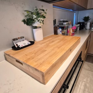 May include: A large, rectangular, light-coloured wooden chopping board with integrated side handles, resting on a kitchen counter. The board features a smooth, multi-piece wood grain construction and a small circular detail on the front edge.
