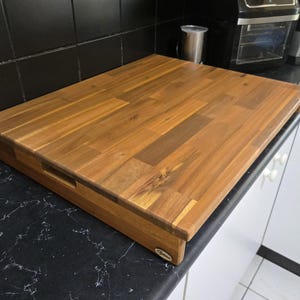 May include: A large, rectangular wooden chopping board with a natural wood finish. The board has a handle on the side and is made of multiple pieces of wood.