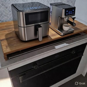 May include: A stainless steel air fryer and espresso machine sit on a wooden surface. The air fryer has a black handle and digital display. The espresso machine has a cup and a digital display.
