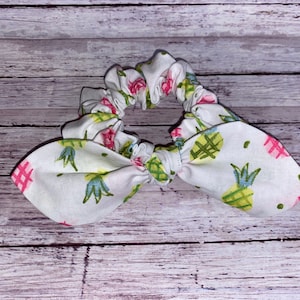 May include: White scrunchie with a bow featuring a pineapple and floral print. The scrunchie has a gathered design and is made of fabric. The bow has a pink hashtag design.