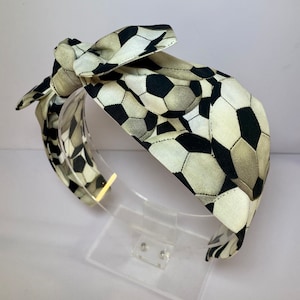 May include: A black and white soccer ball print headband with a bow. The headband is made of fabric and is designed to be worn around the head.