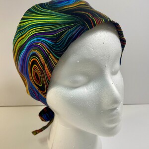 May include: A colorful surgical scrub cap with a swirling pattern of yellow, green, blue, and purple lines on a black background. The cap is tied at the back and is displayed on a white mannequin head.