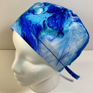May include: A blue and white patterned scrub cap. The cap features a swirling, abstract design in shades of blue, teal, and white. It is tied at the back with a matching fabric tie. The cap is displayed on a white mannequin head.