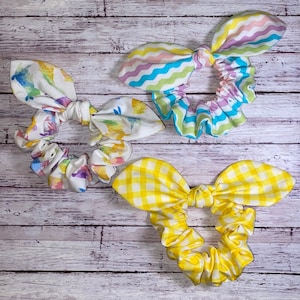 May include: Three colorful hair scrunchies. One is white with a rainbow pattern, one is yellow and white gingham, and one is blue and white striped.
