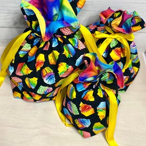 May include: Three drawstring gift bags with a black background covered in colorful cupcake illustrations. Each bag has a rainbow-colored top and is tied with a bright yellow ribbon. The bags are made of fabric and are suitable for gifting.