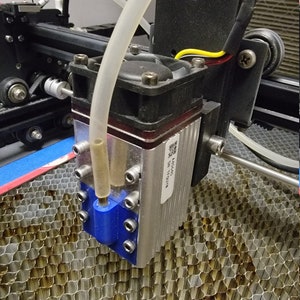 May include: A close-up of a laser cutter head, featuring a blue nozzle and a clear tube. The head is attached to a black frame, with a metal heat sink and a fan. A honeycomb cutting bed is visible beneath.