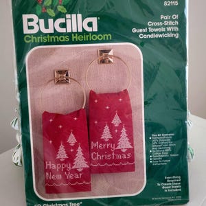 May include: A Bucilla Christmas Heirloom cross-stitch kit for guest towels. The kit includes two red towels with gold-colored rings, featuring white cross-stitched Christmas trees and the phrases "Happy New Year" and "Merry Christmas."