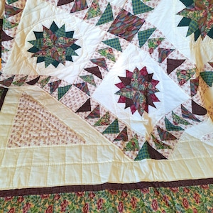 May include: A patchwork quilt featuring a white background with star-shaped motifs in shades of dark green, burgundy, and floral patterns. The design includes triangular fabric segments, a solid white border, and a bottom edge with a floral print.