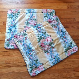 May include: Two square quilted pillow shams featuring a pale yellow background with vertical white stripes. The fabric is decorated with a floral pattern of pink roses, green leaves, and light blue ribbons, finished with a thin blue piped edge.