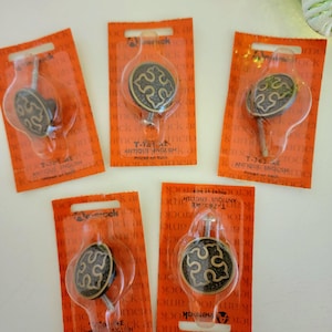 May include: Five round metal cabinet knobs with an antique brass finish and a central cutout design. Each knob is individually sealed in orange Amerock packaging labeled T-761 AE Antique English. The knobs feature a threaded metal screw post.