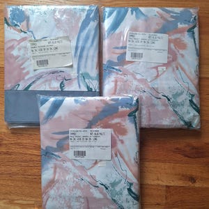 May include: Three packages of window drapes featuring an abstract pattern in shades of blue, pink, and white. Each package includes a white label with printed text detailing the product specifications, including dimensions like 86 inches by 20 inches.