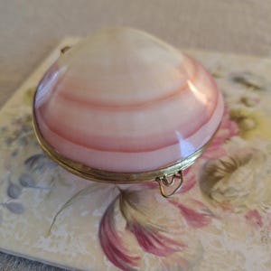 May include: A hinged clam shell trinket box featuring a smooth, polished surface with concentric bands of white and soft pink. The shell is encased in a thin, metallic gold-toned rim with a small, integrated metal loop attached to the front edge.