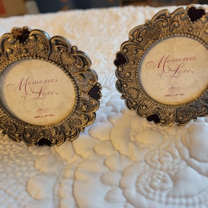 May include: Two ornate, bronze-colored picture frames with a circular design. Each frame has a decorative border with heart-shaped accents and holds a print that reads "Memories of Love." The frames are sized 2 x 2 1/8 inches.