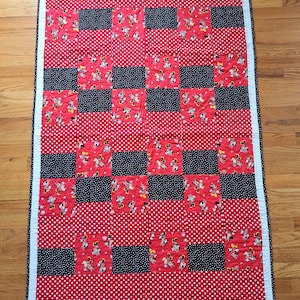 May include: A rectangular quilt featuring a patchwork design of red fabric with white polka dots, black fabric with white polka dots, and red fabric printed with Minnie Mouse characters. The quilt is bordered by a thin white and black trim.