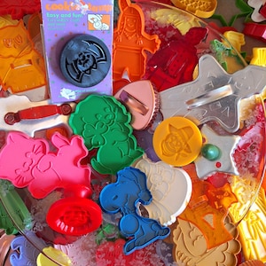 May include: A collection of assorted plastic and metal cookie cutters in various shapes and colors, including a Wilton brand bat cookie stamp in its original packaging, a blue Snoopy character cutter, and various holiday-themed designs.