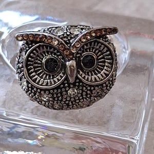May include: A silver-toned ring featuring an owl-shaped centerpiece. The owl has two large, dark circular eyes, a pointed beak, and textured metal feathers. The design is accented with small, clear, and amber-colored rhinestones.