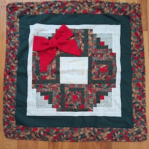 May include: A square quilted fabric mat featuring a log cabin pattern with red, dark green, and cream-colored sections. The border displays a red fabric with a green and gold leaf print. A large, textured red fabric bow is attached to the top left.