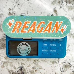 May include: A teal medical device with the name "REAGAN" in orange glitter lettering. The device has a digital display and buttons labeled "ON/OFF", "PRIME", "RUN/PAUSE", "FEED INT", "VOL/TOTAL", "RATE/DOSE", and "CLEAR". A sticker with a bear and the words "MEDICAL MAMA BEAR" is on the device.