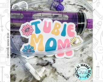 May include: A sticker with the text "Tubie Mom" in colorful letters with flowers around the text. The sticker is on a clear medical tube with a purple syringe and a white connector.