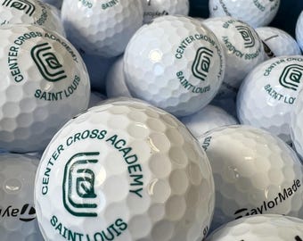 May include: A collection of white golf balls with dimpled surfaces. Some balls feature the text "TaylorMade" and numbers, while others display "CENTER CROSS ACADEMY SAINT LOUIS" in green lettering.