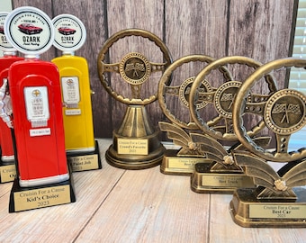 May include: Five gold-colored trophies with different designs. The trophies are on a wooden surface. The trophies are for a car show called "Cruisin' For a Cause".