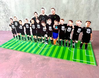 Könnte beinhalten: Eine transparente Acryl-Ausstellung mit einer Fußballmannschaft und ihren Trainern auf einem grün-weißen Feld. Das Team trägt schwarze Trikots mit einem Fußballlogo. Die Trainer tragen schwarze Hemden. Die Ausstellung befindet sich auf einer rosa Oberfläche.