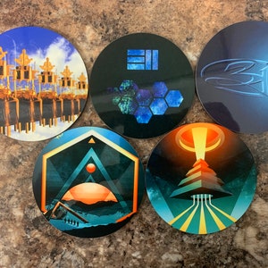 May include: Five round coasters with different designs. The coasters are made of a hard material and have a glossy finish. The designs include a geometric pattern, a cityscape, a stylized logo, a futuristic scene, and a honeycomb pattern.