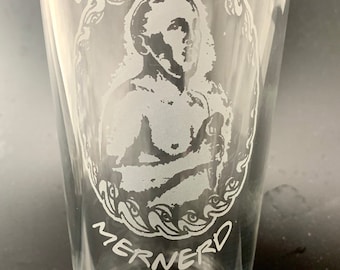 Könnte beinhalten: Klares Bierglas mit geätztem Design. Das Design zeigt ein Porträt einer Person in einem dekorativen Rahmen mit den Worten "ERMAGHERD" und "MERNERD". Das Glas hat eine konische Form.